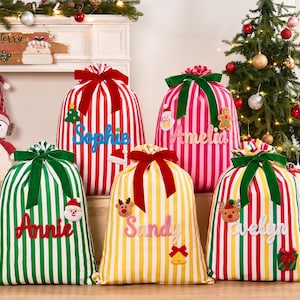 Pode incluir: Cinco sacos de presente de Natal em tecido com listras verticais em vermelho, verde, rosa e amarelo. Cada saco é amarrado com uma fita de veludo e personalizado com nomes em letras de feltro, incluindo Sophie, Amelia, Annie, Sandy e Evelyn.