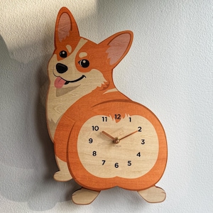 May include: A wall clock shaped like a Corgi dog with orange and cream wood grain patterns. The clock face is integrated into the dog's rear, featuring black numerals from 1 to 12, two thin gold-colored hands, and the brand name YOHUS printed.