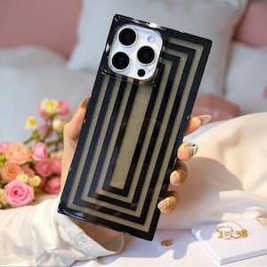 Peut inclure: Une coque de smartphone rectangulaire dotée d'une base beige translucide ornée d'une série de cadres rectangulaires noirs concentriques. La coque présente une découpe carrée pour l'appareil photo en haut à gauche, révélant trois objectifs.