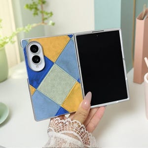 May include: A foldable smartphone case featuring a geometric patchwork design in shades of mustard yellow, light blue, dark blue, and sage green. The back panel includes a raised white camera housing with two circular lenses and a silver frame.