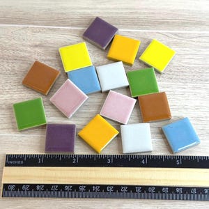 Peut inclure: Une collection de dix-sept petits carreaux de céramique carrés et multicolores dans des tons de jaune, vert, bleu, violet, rose, marron et blanc, disposés au-dessus d'une règle noire indiquant des mesures en centimètres pour l'échelle.