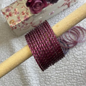 May include: A set of deep purple bangles with a textured, faceted surface and small gold-coloured accents, arranged in a stack on a cylindrical beige display stand. The bangles feature a repeating pattern of raised, angled segments.