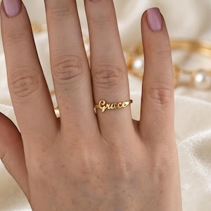 May include: A gold-toned ring featuring the name Grace in a cursive script font. The ring is thin and polished, with the lettering integrated directly into the band. The design is simple and metallic, showcasing a smooth finish on the surface.