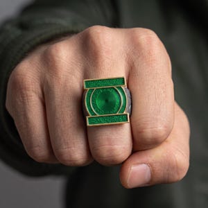 Op de afbeelding: Een ring met een opvallend groen cirkelvormig embleem met een centraal spiraalpatroon, omlijst door goudkleurige horizontale balken met een getextureerd oppervlak. De ringband heeft een donkere, ruwe en onregelmatige afwerking, in contrast met het groen.