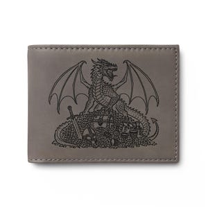 May include: A grey bi-fold wallet featuring a black laser-engraved illustration of a dragon perched atop a pile of treasure, including a sword, a helmet, goblets, and a chest, with visible stitching along the edges of the leather surface.