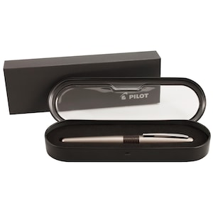 Puede incluir: Una pluma estilográfica color champán con una sección de agarre texturizada en marrón oscuro y un clip plateado, dentro de un estuche de presentación ovalado negro con tapa transparente. El logo de Pilot está impreso en blanco en la tapa.