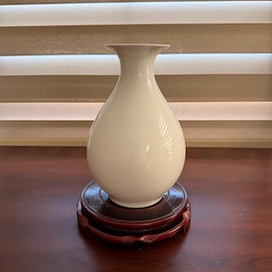 Puede incluir: Un jarrón de cerámica blanca lisa con cuerpo bulboso y borde acampanado. El jarrón está colocado sobre una base de exhibición circular de madera de color marrón rojizo oscuro con detalles tallados, acabado brillante y diseño simple.