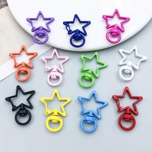 Puede incluir: Un conjunto de doce cierres de mosquetón con forma de estrella en varios colores, incluyendo morado, azul oscuro, rosa, naranja, rosa claro, verde, blanco, negro, amarillo, azul claro y rojo, cada uno con base giratoria y anilla.