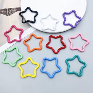 Puede incluir: Una colección de doce anillas metálicas con cierre de resorte en forma de estrella en varios colores, incluyendo negro, blanco, morado, rosa, naranja, rojo, rosa claro, verde claro, amarillo, azul y verde azulado, con un pequeño mecanismo.
