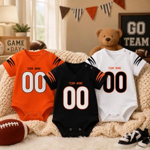 May include: Three short-sleeved baby bodysuits in orange, black, and white. Each features a V-neck, snap closures at the shoulder and crotch, and tiger-stripe patterns on the sleeves. The text "TEAM NAME" and the number "00" are printed on the front.