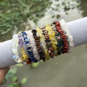 May include: A collection of stretch bracelets made from various polished gemstone chips. The bracelets feature a wide array of colors including white, purple, blue, pink, brown, yellow, green, and red, arranged in a row on a gray cylindrical display.