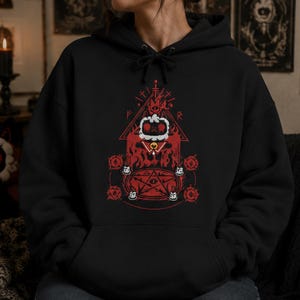 Pode incluir: Uma camisola com capuz preta com uma impressão gráfica em vermelho e branco. O design apresenta uma personagem central com cabeça branca, túnica vermelha e coleira com sino, posicionada sobre um pentagrama rodeado por quatro velas pequenas.