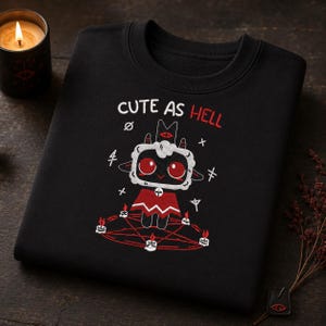 May include: A black sweatshirt featuring an embroidered graphic of a cartoon lamb with large red eyes, wearing a red cape. The text CUTE AS HELL is printed above the lamb, which stands inside a red pentagram surrounded by small white candles.