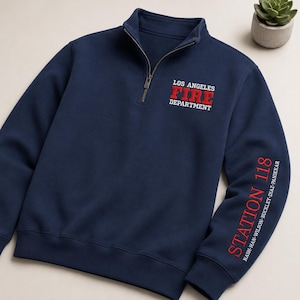 Puede incluir: Una sudadera azul marino con cremallera de un cuarto, con el texto bordado en blanco y rojo LOS ANGELES FIRE DEPARTMENT en el pecho y STATION 118 con los nombres NASH-HAN-WILSON-BUCKLEY-DIAZ-PANIKKAR impresos en rojo y blanco en la manga.