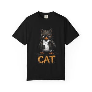 May include: A black short-sleeve t-shirt featuring a graphic of a stylized owl wearing a leather jacket and a mask. Below the owl, the word CAT is printed in a distressed, orange, stencil-style font. The design is centered on the chest area.