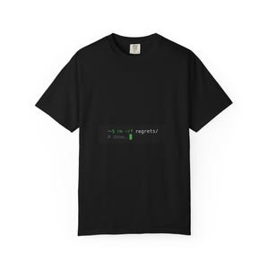 Op de afbeelding: Een zwart t-shirt met korte mouwen en een grafische print van een computerterminal-opdrachtregel. De tekst in groen lettertype luidt: ~$ rm -rf regrets/ gevolgd door # done. met een klein groen cursorblokje aan het einde.