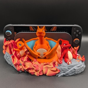 May include: A gaming console stand featuring a central orange Charizard figure with blue-lined wings, flanked by a small orange Charmander and a red Charmeleon. The base is sculpted to resemble gray rock with swirling red and orange flames.