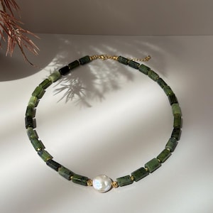 May include: A necklace featuring a series of rectangular dark green stone beads interspersed with small gold-toned metal spacers, centered with a single round white pearl, finished with a gold-toned metal clasp and chain extender.