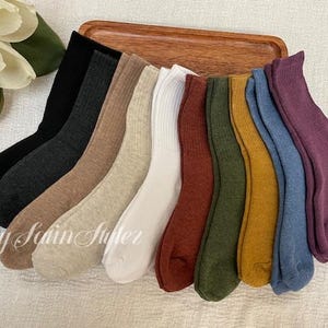 May include: A collection of ten ribbed crew socks in various colors including black, charcoal, beige, cream, white, rust, olive green, mustard yellow, blue, and plum, arranged in a fan shape with the text By Scin Styler visible on the left.