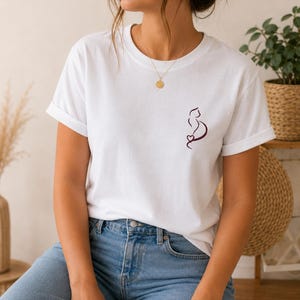 Puede incluir: Una camiseta blanca de manga corta con un gráfico lineal minimalista de color morado oscuro que muestra la silueta de un gato con un pequeño corazón en el pecho izquierdo, combinada con vaqueros azul claro y un collar con colgante dorado.