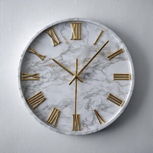 May include: A round wall clock featuring a white marble face with grey veining. The clock has polished gold-colored Roman numerals for hour markers and matching gold-colored hour, minute, and second hands centered on the dial.