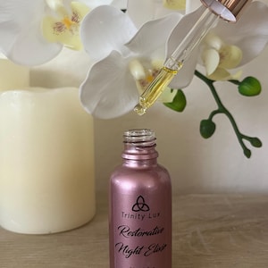 May include: A metallic rose gold glass bottle of Trinity Lux Restorative Night Elixir with a 30mL/1 fl oz capacity. The bottle features a dropper cap with a rose gold collar and a clear glass pipette dispensing a golden yellow liquid oil.