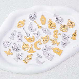 Peut inclure: Une collection de petites breloques en métal argenté et doré représentant divers motifs sur le thème d'Halloween, notamment des crânes, citrouilles, toiles d'araignée, cercueils, chauves-souris, lunes, étoiles, bougies, lanternes, chapeaux de sorcière et masques.