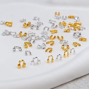 Könnte beinhalten: Eine Ansammlung kleiner, U-förmiger Quetschperlenkappen aus Metall in Gold- und Silbertönen. Diese Schmuckzubehörteile haben ein hohles, abgerundetes Design mit zwei parallelen zylindrischen Enden, um Knoten oder Quetschperlen zu verdecken.