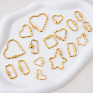 May include: A collection of gold-toned metal spring gate rings in various shapes including hearts, stars, rectangles, and ovals. Each piece features a textured or smooth screw-lock mechanism for opening and closing, designed for jewelry or accessory use.