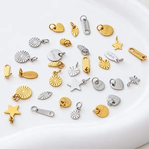 May include: A collection of small metal jewellery charms in gold and silver tones, featuring various shapes including stars, hearts, circles, and elongated rectangles. Many pieces are stamped with the text S.steel and have jump rings attached.