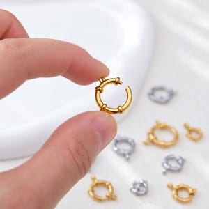 May include: A gold-toned circular spring ring clasp with a small lever mechanism for jewelry making. The clasp features a smooth, polished finish and a small loop attachment point. Several additional spring ring clasps in gold and silver tones are scattered nearby.