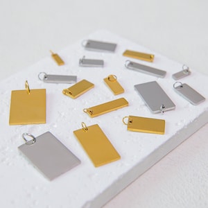 Könnte beinhalten: Eine Sammlung rechteckiger Metallanhänger in Gold- und Silbertönen, die jeweils mit einem kleinen runden Biegering an der Oberseite versehen sind. Die Anhänger variieren in der Größe und sind verstreut angeordnet, mit glatten, flachen Oberflächen.