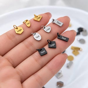 Könnte beinhalten: Eine Sammlung kleiner Metallanhänger in Gold-, Silber- und Schwarztönen mit verschiedenen geometrischen Formen wie Sechsecken, Herzen, Rechtecken und Kreisen. Jeder Anhänger verfügt über einen Biegering und ist mit dem Wort Text oder Logo graviert.