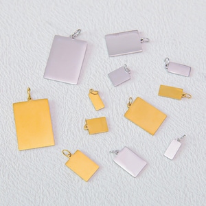 May include: A collection of rectangular metal charms in various sizes, featuring both gold-tone and silver-tone finishes. Each charm includes a small circular jump ring at the top for attachment, with smooth, flat surfaces and rounded corners.