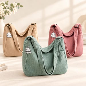 May include: Three corduroy hobo bags in beige, dusty pink, and sage green. Each bag features a wide shoulder strap, a side pocket, and a small white dog-shaped charm attached to the strap with a silver-toned metal chain.