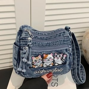 Könnte beinhalten: Eine blaue Umhängetasche aus Jeansstoff mit drei gestickten Katzen auf der Vordertasche. Die Tasche verfügt über mehrere Reißverschlussfächer mit silberfarbenen Beschlägen, Reißverschlussanhängern aus Jeansstoff und dem Schriftzug aishangtao.