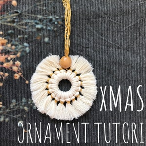 May include: White macrame Christmas ornament with a gold thread design and a wooden bead. The ornament is hanging from a gold cord. The text "XMAS ORNAMENT TUTORIAL" is written below the ornament.
