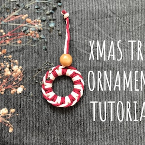 May include: A red and white striped yarn Christmas tree ornament with a wooden bead at the top. The ornament is hanging from a white string. The text "XMAS TREE ORNAMENT TUTORIAL" is written in white on a grey background.