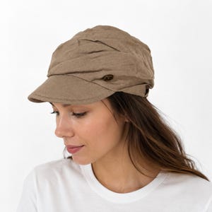 May include: A light brown newsboy cap featuring a gathered, pleated crown design and a short, stiff brim. The cap is accented with a small, dark brown circular button detail on the side, providing a textured appearance to the fabric surface.