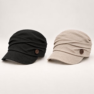 May include: Two newsboy caps, one in black and one in beige, featuring a textured fabric, a rounded crown with horizontal pleated detailing, a short stiff brim, and a single brown four-hole button accent on the side of each cap.