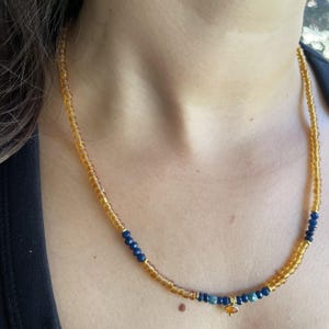 May include: A beaded necklace featuring a long strand of translucent amber-coloured glass beads, accented with sections of small, faceted dark blue beads. A single, small, oval-shaped amber-coloured charm hangs from the centre of the necklace.
