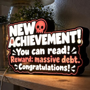 May include: A rectangular LED sign with a black frame and red glowing edges. The text reads: NEW ACHIEVEMENT! with a skull icon, You can read!, Reward: massive debt., Congratulations!. The sign is illuminated and features white lettering.