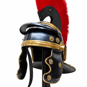 Könnte beinhalten: Ein Helm im römischen Stil mit einer schwarzen Metallkuppel und Wangenklappen, verziert mit goldfarbenen Metallrändern und kreisförmigen Akzenten. Der Helm ist mit einem hohen, leuchtend roten Kamm auf einer schwarzen Halterung versehen.