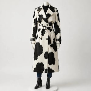 May include: A long, double-breasted trench coat featuring a black and white cow-print pattern. The coat includes a notched collar, a matching tie belt with a black buckle, shoulder epaulets, and black leather-like straps on the sleeve cuffs.