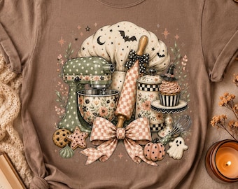 May include: A brown short-sleeve t-shirt featuring a graphic print of a green stand mixer, a checkered rolling pin, a canister, a cupcake with a witch hat, a whisk, cookies, a ghost, and a pumpkin, all tied together with a large checkered bow.