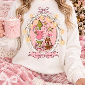 Può includere: Una felpa bianca a maniche lunghe con una grafica centrale all'interno di una cornice ovale. Il disegno mostra un personaggio verde in abito rosa che tiene una piccola figura, un cane marrone con cappello da Babbo Natale, un albero rosa e luci.