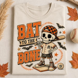May include: A beige t-shirt featuring a graphic of a skeleton wearing a checkered baseball cap and uniform, holding a bat. The text reads BAT TO THE BONE. The design includes orange and black accents, small bats, stars, and a baseball with a skull.