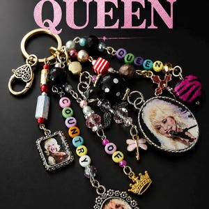 Peut inclure: Un porte-clés composé de plusieurs chaînes de perles et de breloques, incluant trois cadres ovales et carrés avec des portraits d'une chanteuse blonde, des perles lettres formant COUNTRY QUEEN, une breloque couronne et un fermoir en cœur.