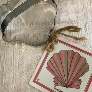 May include: A metal seashell-shaped cookie cutter with a handle, next to a tag featuring a seashell illustration in pink and brown, with the word "Seashell". The tag is tied with twine.