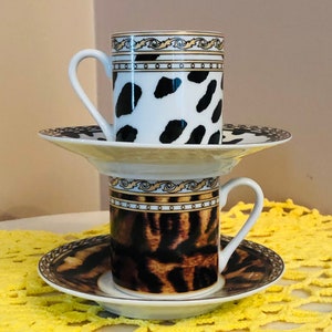 Op de afbeelding: Twee gestapelde espressokopjes en schotels. De bovenste kop heeft een witte achtergrond met zwarte vlekken, terwijl de onderste kop een bruine en zwarte dierenprint heeft. Beide kopjes hebben gouden randen en witte oren. De schotels hebben bijpassende patronen.